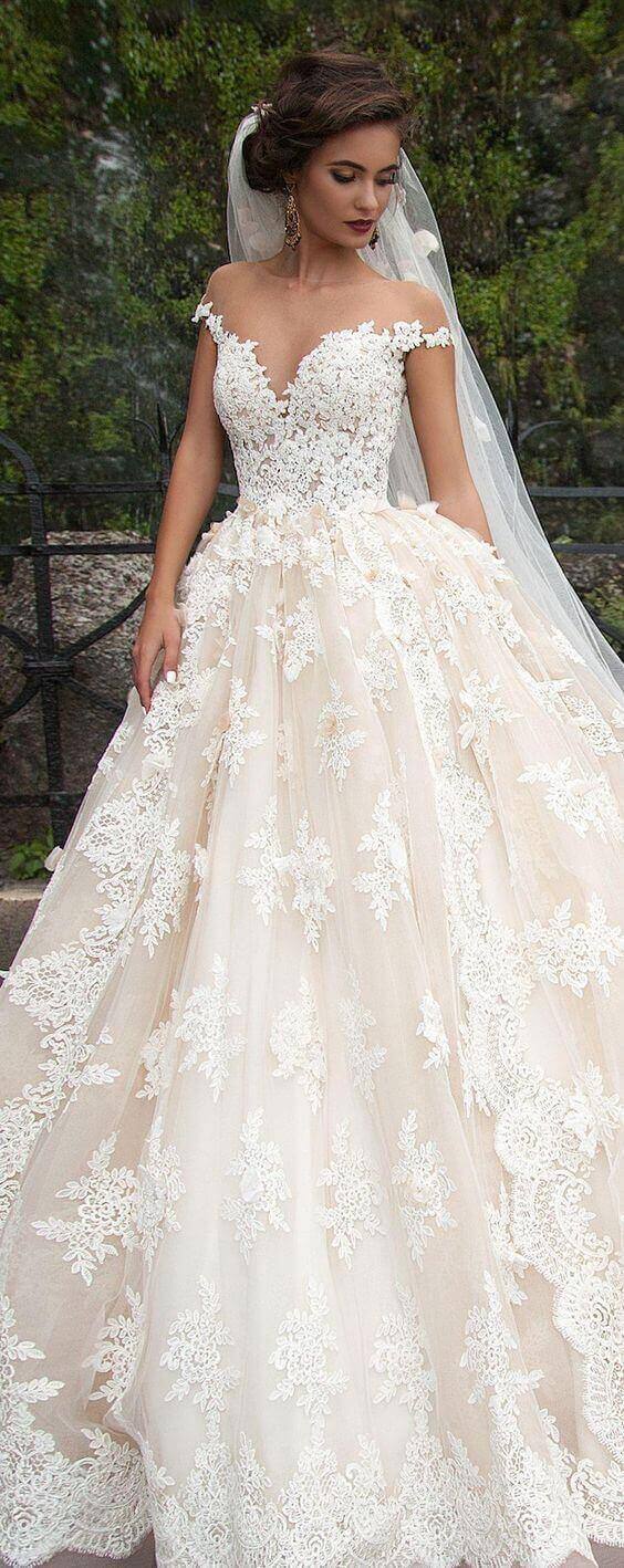 47 perfect bridal gown ideas - find yours 3 A bride in an ivory-champagne ballgown with off-the-shoulder lace stands in a garden, showcasing intricate floral appliqués and a cathedral veil.