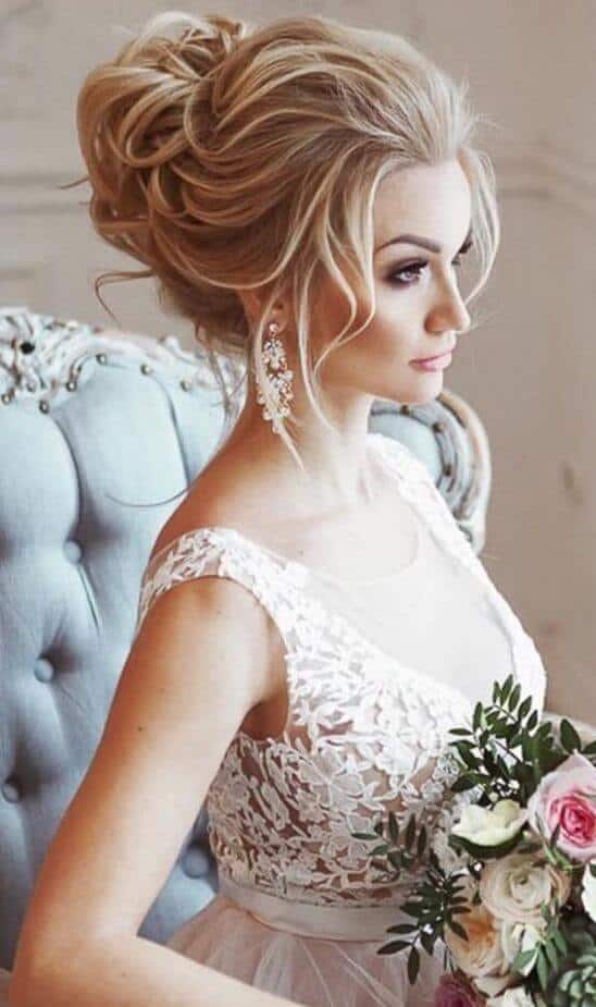39 popular wedding hairstyles for brides to make your big day perfect 20 Bride in a romantic bridal portrait, showcasing a voluminous updo, lace gown, and pastel bouquet in a serene setting.