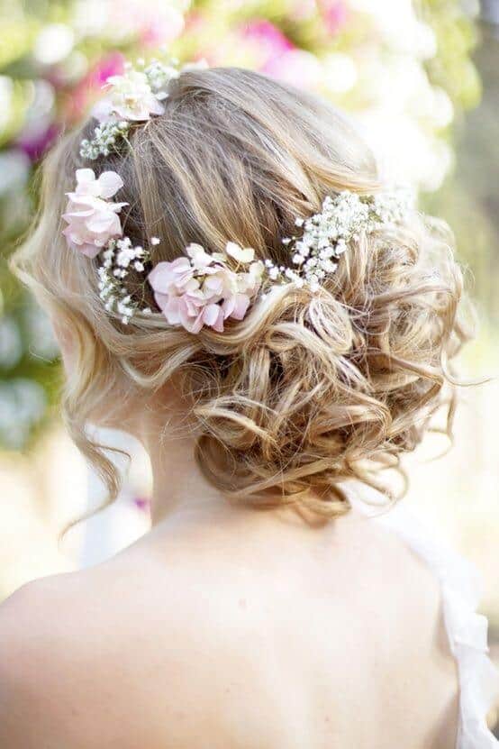 Bridal low curly updo adorned with blush pink flowers and baby's breath, set in a garden, exuding a romantic wedding vibe.