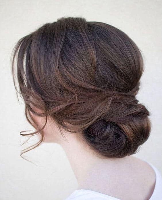 Romantic low chignon bridal updo with soft face-framing tendrils, showcasing brunette hair with cocoa highlights against a neutral backdrop.