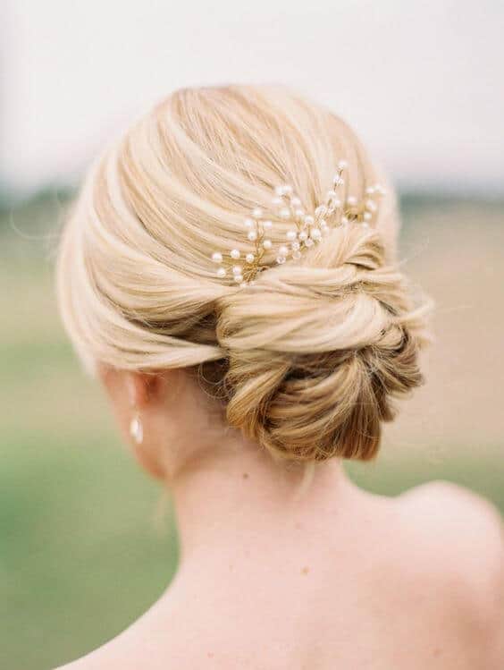 Back view of a romantic low chignon featuring soft twists and a pearl sprig, set against a blurred outdoor background.