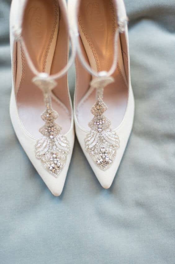 Ivory t-strap wedding shoes with art deco crystal appliques, elegantly arranged on soft blue fabric, viewed from above.