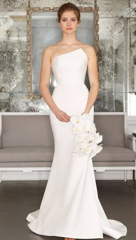 47 perfect bridal gown ideas - find yours 25 Poised bride in a modern lounge, wearing a one-shoulder satin wedding gown with an asymmetrical neckline, holding a cascading white orchid bouquet.
