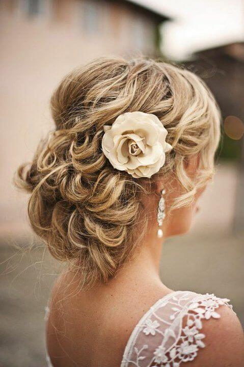 Romantic wedding updo featuring a low chignon styled with loose barrel curls. An ivory rose accessory complements the lace-appliquéd gown.