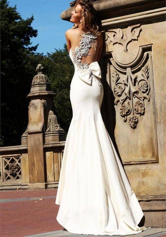 47 perfect bridal gown ideas - find yours 35 Backless mermaid wedding dress with lace appliqué and statement bow, modeled by a bride on a sunlit historic stone terrace.