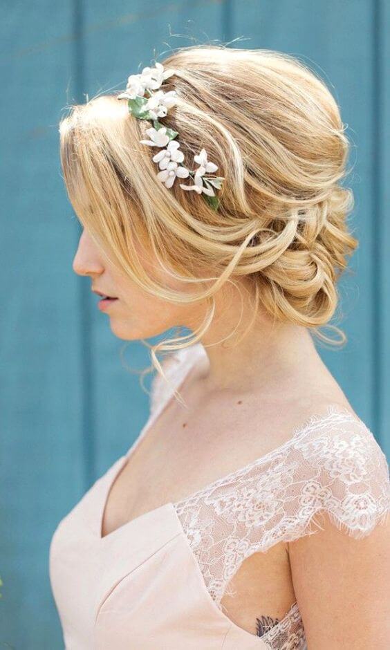 Bridal portrait featuring a romantic low chignon with face-framing tendrils, adorned with a floral headband, against a dusty-blue backdrop.