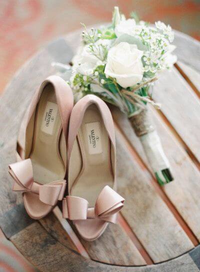 Blush pink peep-toe heels with bow accents alongside a bridal bouquet of ivory roses and greenery on a rustic wooden table.