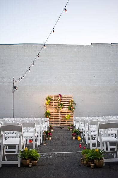 31 lovely and beautiful cheap wedding venues ideas 21 Urban outdoor wedding setup in a minimalist courtyard, featuring a rustic pallet altar adorned with greenery and vibrant flowers under bistro lights.