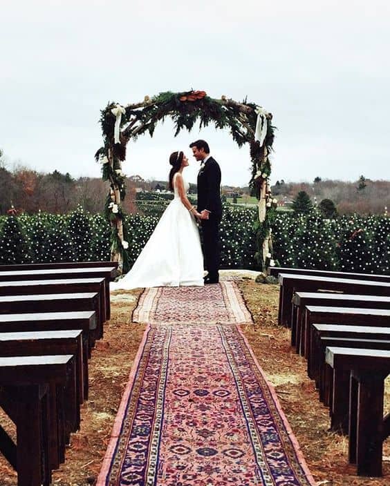 31 lovely and beautiful cheap wedding venues ideas 27 A bride and groom stand hand-in-hand beneath a wooden arch adorned with evergreen garlands and white blooms, surrounded by vintage rugs and wooden benches.