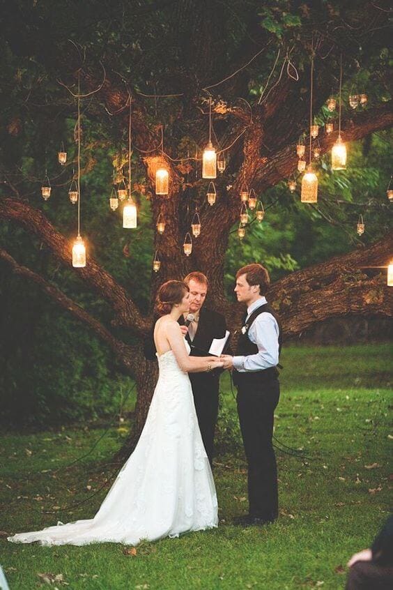 31 lovely and beautiful cheap wedding venues ideas 5 Bride and groom exchange vows under a large tree at dusk, surrounded by hanging mason-jar candles creating a warm, romantic glow.