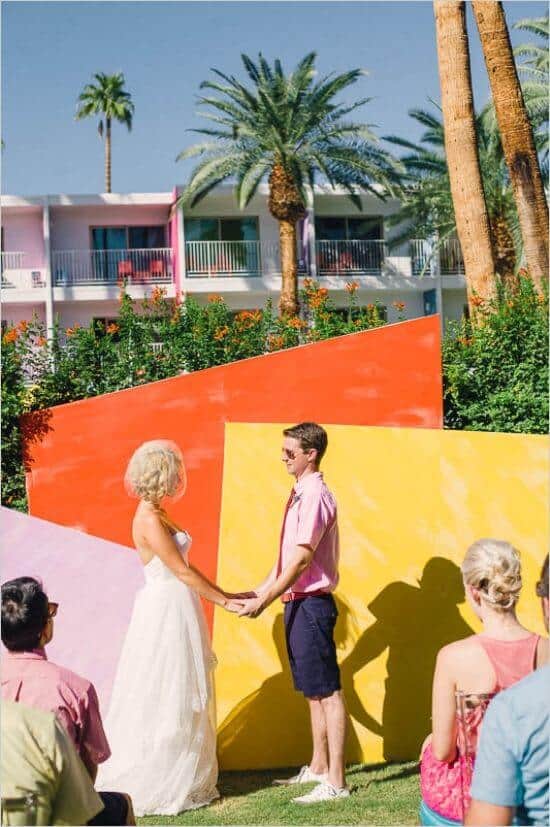 31 lovely and beautiful cheap wedding venues ideas 33 Outdoor cheap wedding ceremony with a bride in a white gown and partner in pink shirt before colorful geometric panels, surrounded by palm trees.