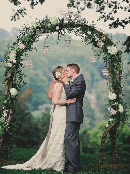 31 lovely and beautiful cheap wedding venues ideas 9 Bride and groom share a romantic first kiss beneath a rustic floral arch, surrounded by lush greenery in a serene garden setting.