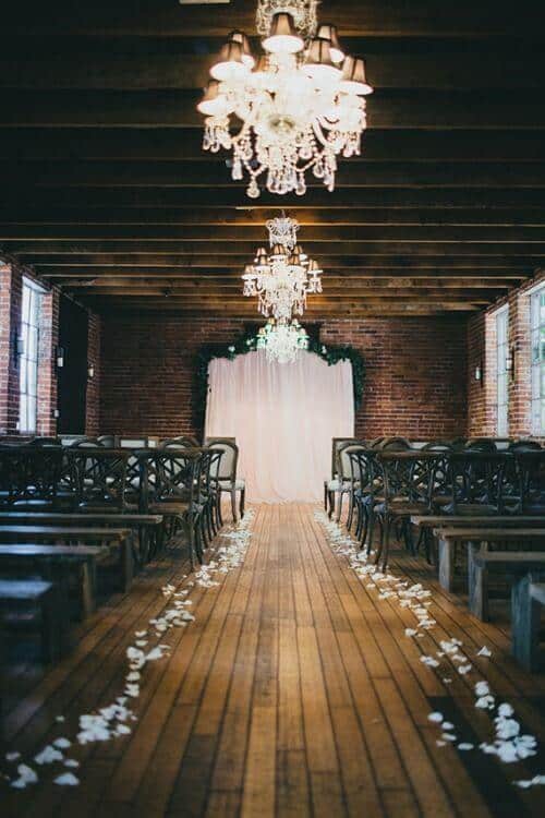 Rustic indoor wedding venue featuring a petal-lined wooden aisle, crystal chandeliers, and an ivory draped backdrop framed with greenery.