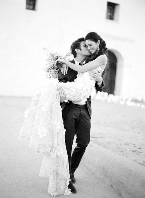 50 bride and groom photo ideas to save to posterity 13 Groom carries his smiling bride in a lace gown with a long train, kissing her cheek in front of a white-stucco building—an intimate, black-and-white wedding portrait.