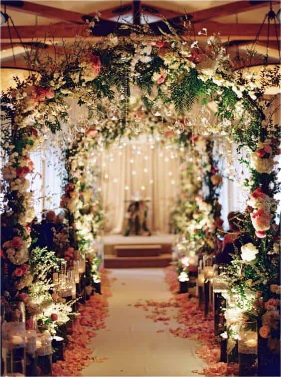 Candlelit floral tunnel framing a wedding aisle, adorned with blush and ivory blooms, glowing lights, and a romantic altar backdrop.