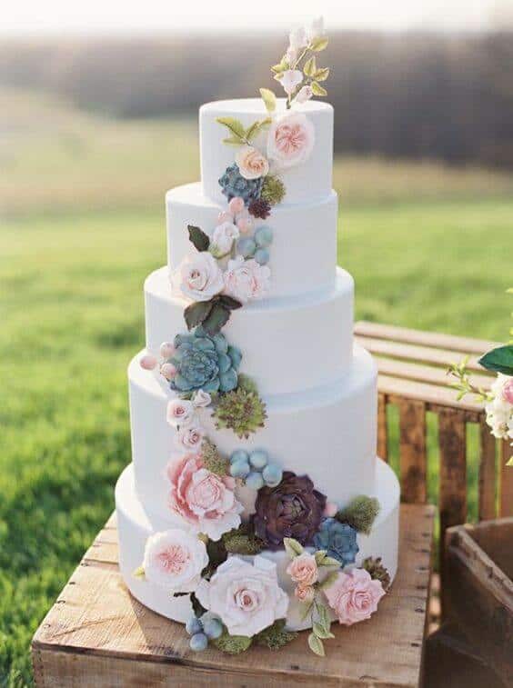 41 best wedding cake designs ideas for your big day 14 Five-tier white fondant wedding cake with cascading blush roses, pastel succulents, and dusty blue berries, staged on rustic crates in a meadow.