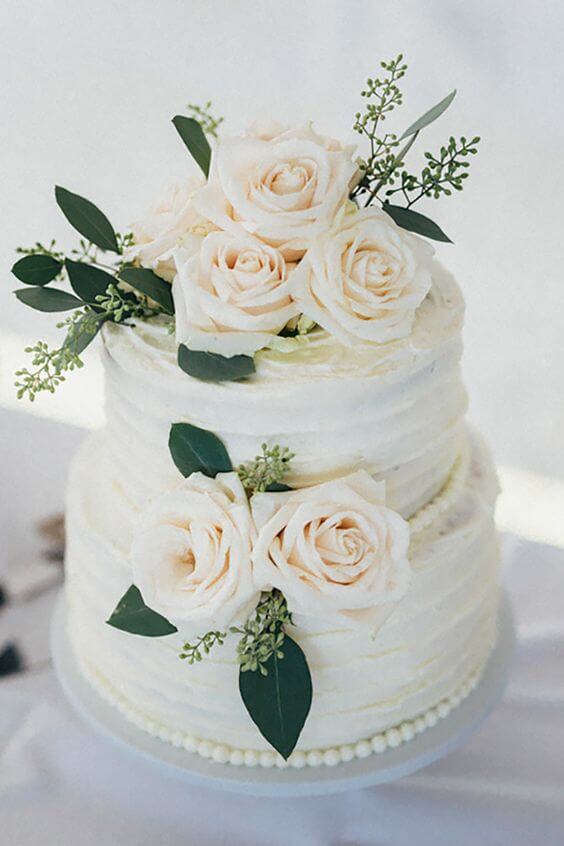41 best wedding cake designs ideas for your big day 16 Two-tier ivory buttercream wedding cake adorned with ivory roses and eucalyptus, displayed on a white pedestal stand.