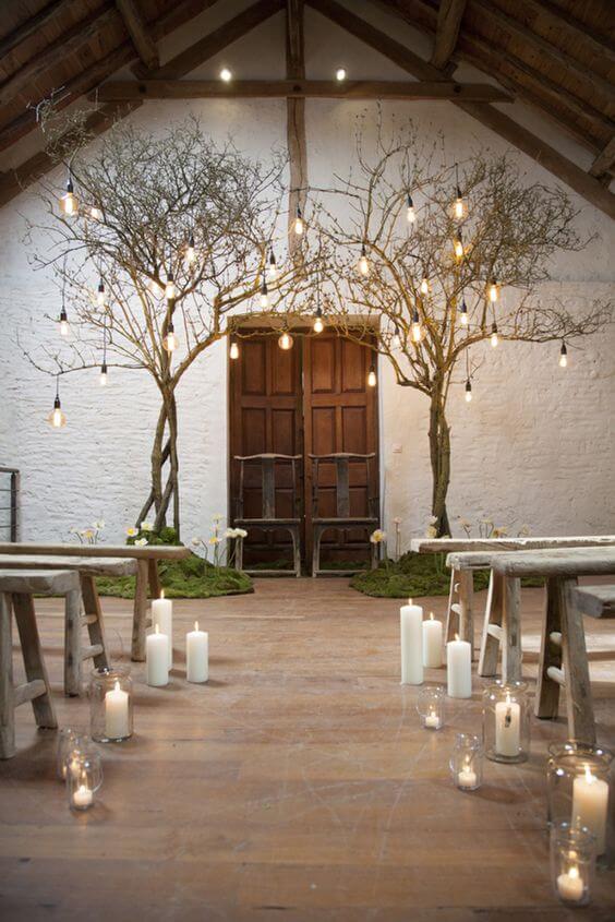Candlelit indoor wedding ceremony in a rustic barn, featuring a wooden aisle, leafless trees, and warm edison bulb lighting.