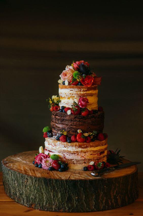 41 best wedding cake designs ideas for your big day 18 Rustic semi-naked wedding cake with three tiers of vanilla and chocolate sponges, adorned with fresh berries and blooms, on a wood slab.