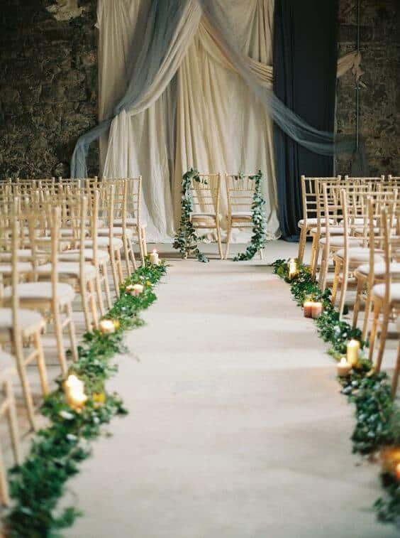 Stone-walled venue with gold chiavari chairs facing a draped altar, surrounded by greenery and candlelight for an intimate wedding setup.