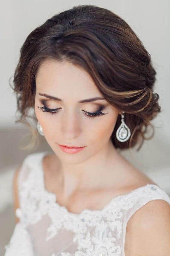 Close-up bridal portrait featuring soft glam makeup with taupe-bronze eyeshadow, peach-coral lips, and an ivory lace illusion neckline.