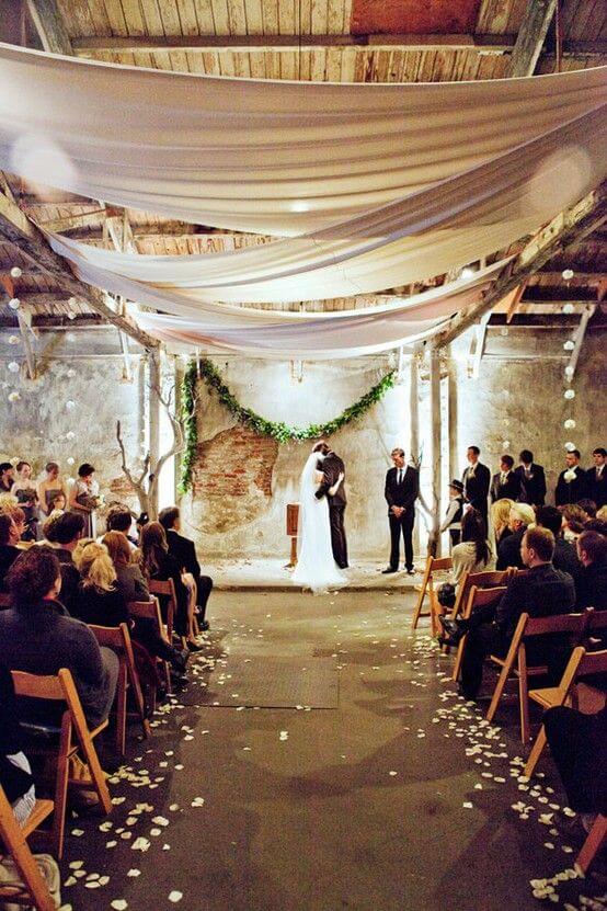 Romantic indoor wedding ceremony in a rustic warehouse. Couple kissing under a chiffon canopy, surrounded by greenery and soft lighting.