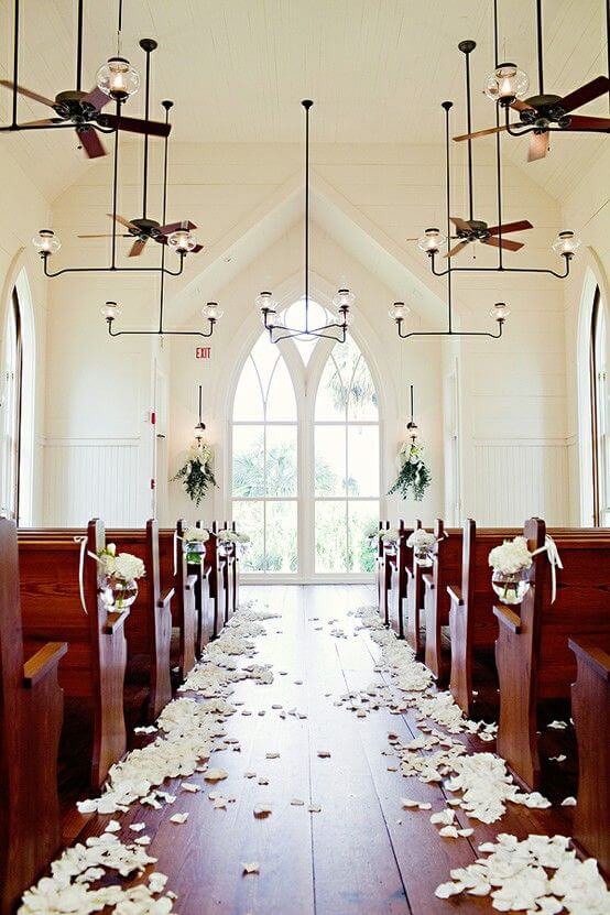 Bright chapel with petal-lined aisle, gothic windows, wooden pews, and glass jar florals, creating a serene wedding venue atmosphere.