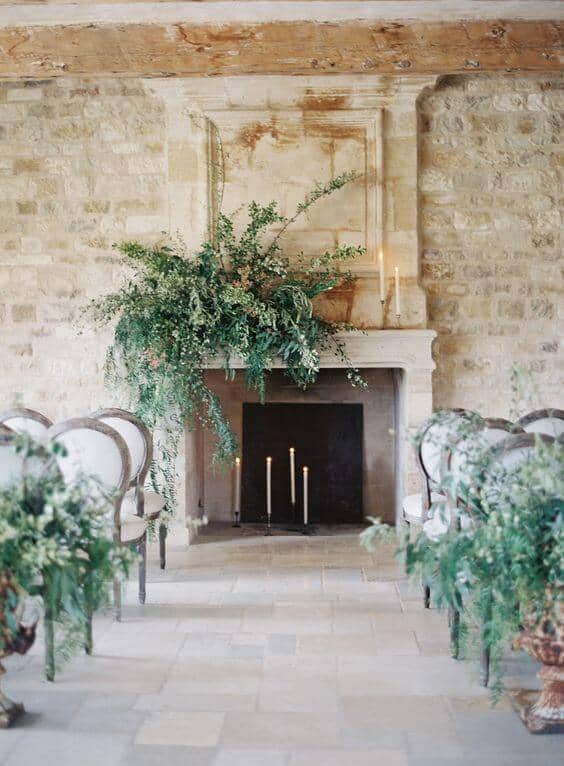 Intimate indoor wedding setup featuring a limestone fireplace, lush greenery mantel, and elegant chairs in a rustic manor.