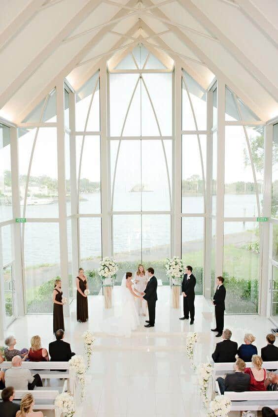 A luminous indoor wedding in a glass chapel, with the couple on a white platform, framed by floral arrangements, and a serene waterfront view.