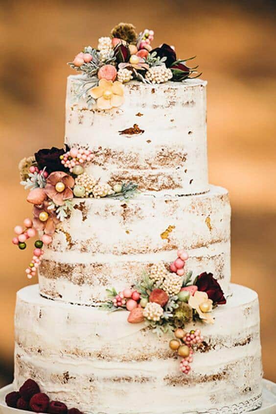 41 best wedding cake designs ideas for your big day 25 Three-tier semi-naked wedding cake adorned with blush and peach berries, ivory blossoms, and gold leaf, set against warm amber bokeh.