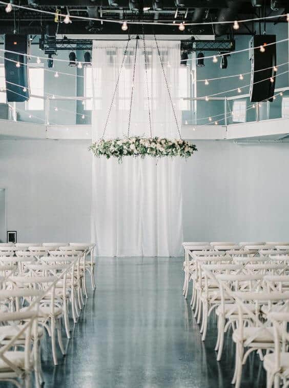 Minimalist indoor wedding venue featuring white cross-back chairs, floral chandelier, and soft drapery, creating an airy atmosphere.