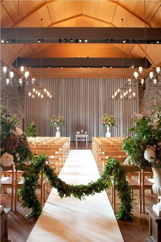 Refined indoor wedding venue setup featuring a timber-vaulted ceiling, ivory aisle runner, greenery garland, and elegant floral arrangements.
