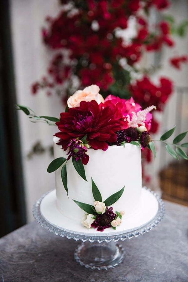 41 best wedding cake designs ideas for your big day 29 White buttercream wedding cake on a glass pedestal, adorned with deep burgundy and blush florals, evoking a romantic garden vibe.