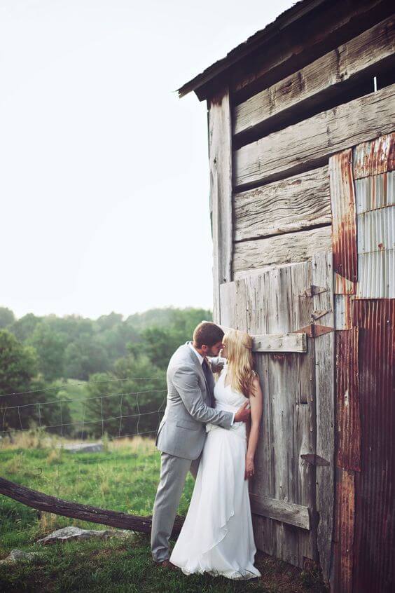 50 bride and groom photo ideas to save to posterity 29 Bride and groom sharing a kiss by a weathered barn door during a rustic countryside wedding, framed by natural textures and soft daylight.