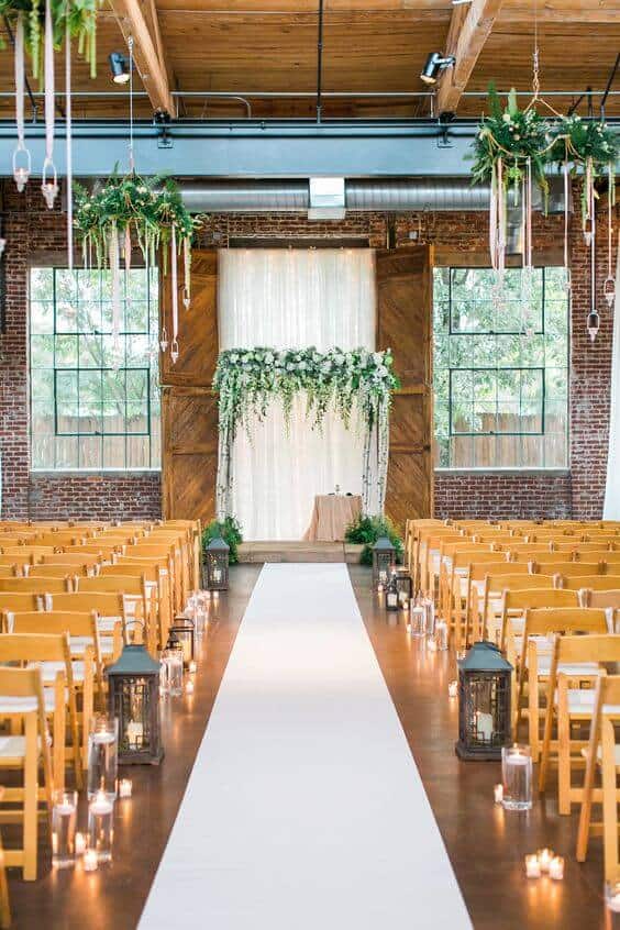 Industrial loft wedding venue featuring a white aisle runner, rustic altar with greenery, and warm candlelight ambiance.