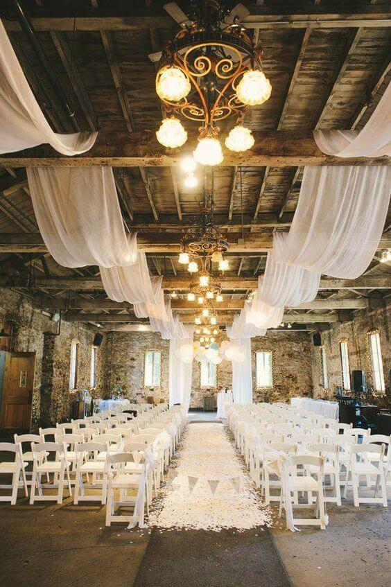 Rustic barn wedding ceremony aisle featuring white drapery, vintage chandeliers, and petal-strewn path leading to a fabric-draped altar.