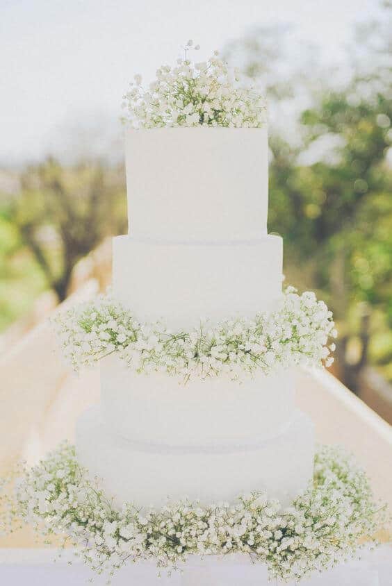 41 best wedding cake designs ideas for your big day 5 Elegant four-tier wedding cake with smooth white fondant and baby’s breath accents, displayed in a sunlit garden setting.