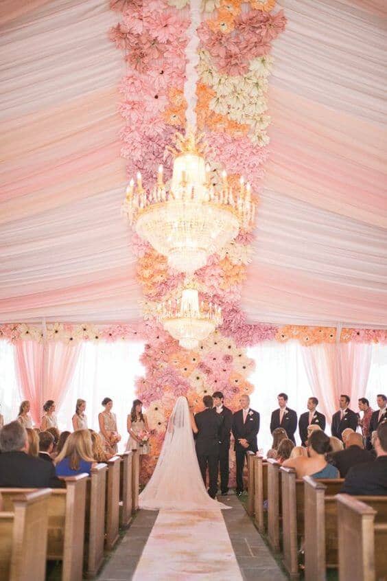 Romantic indoor tented wedding ceremony with blush drapery, ombré floral installation, and crystal chandeliers, framing a couple at the altar.