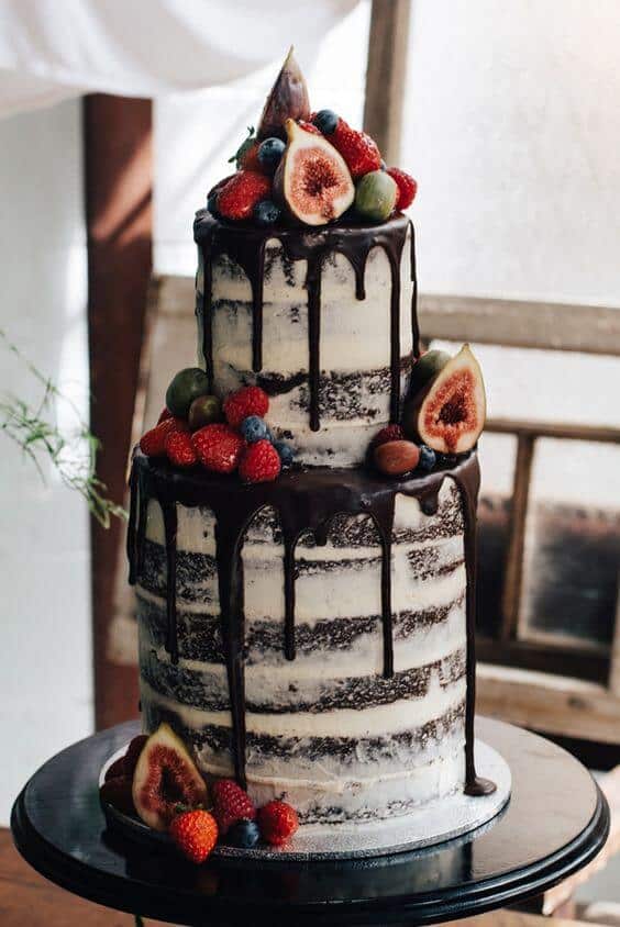 41 best wedding cake designs ideas for your big day 33 Two-tier semi-naked chocolate drip wedding cake adorned with fresh figs, raspberries, blueberries, and grapes on a black pedestal in a rustic setting.