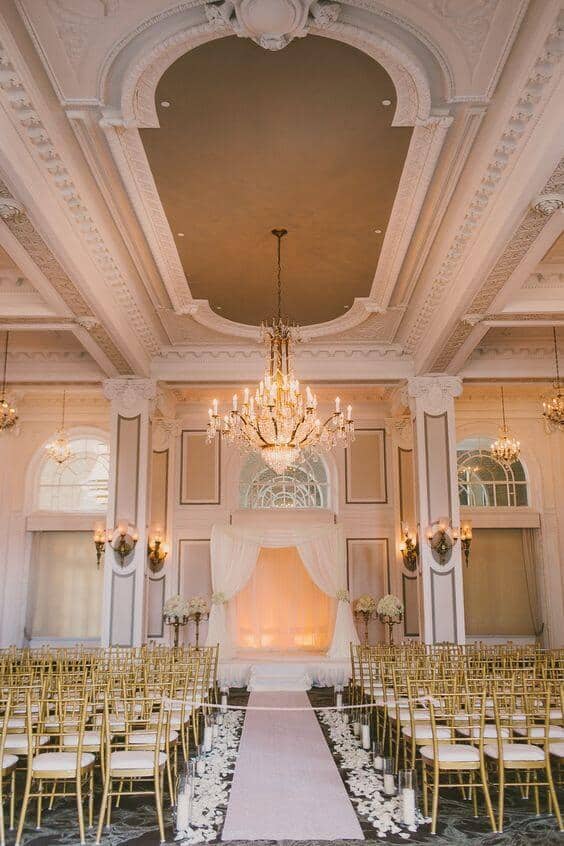 Luxurious indoor wedding venue featuring a grand ballroom setup with gold chiavari chairs, an ivory aisle runner, and a draped ceremony arch.