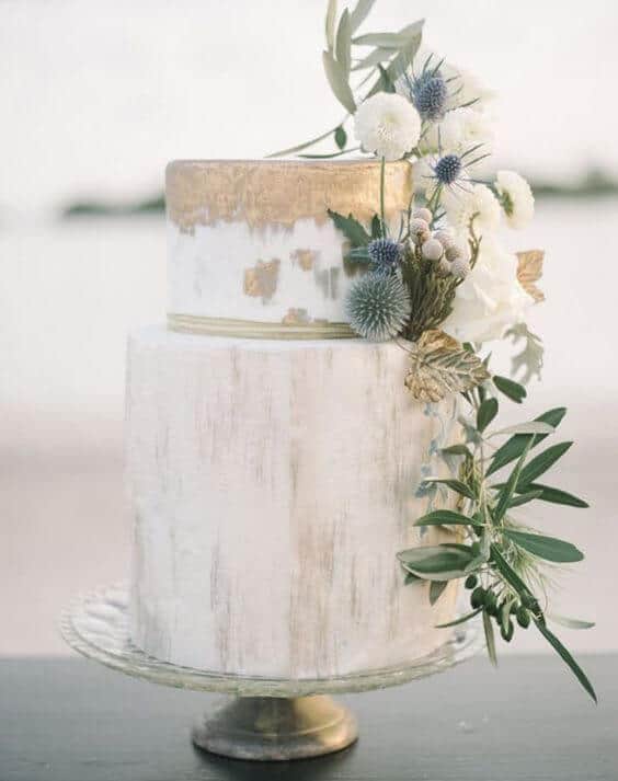 41 best wedding cake designs ideas for your big day 37 {{post_main_keyword}} featuring a two-tier wedding cake with a gold-leaf top tier, adorned with blue thistles and white blooms on a glass pedestal.