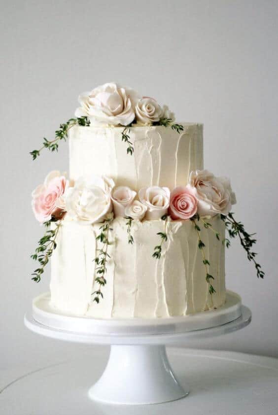 41 best wedding cake designs ideas for your big day 6 Two-tier ivory buttercream wedding cake adorned with soft blush roses and trailing greenery, set against a light gray backdrop.