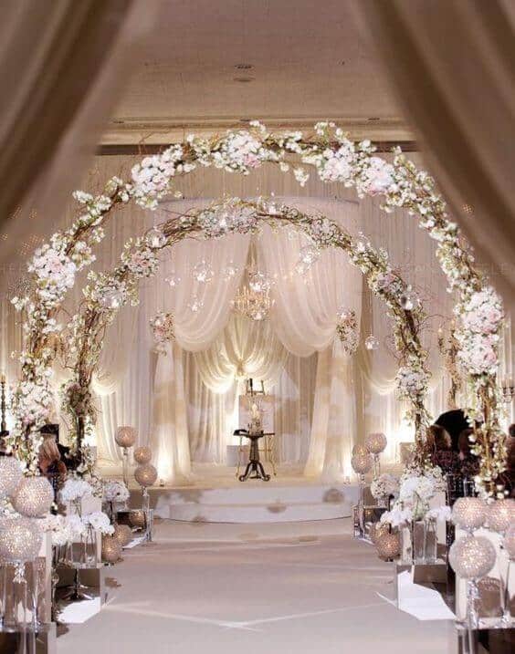 Ethereal indoor wedding venue with a soft-ivory aisle, floral arches, and a multi-tier wedding cake on a raised platform, surrounded by crystal chandeliers.