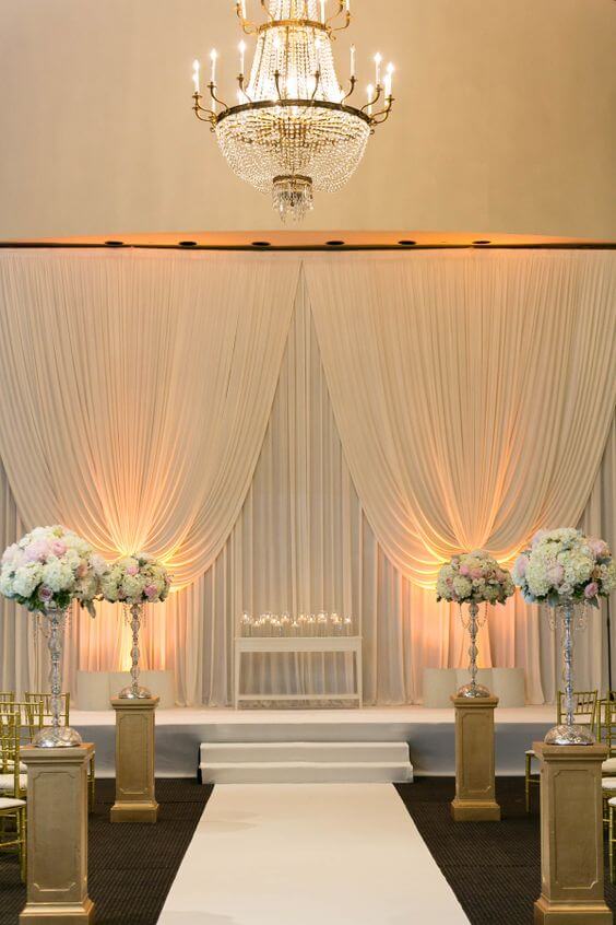 Elegant ivory-draped altar in a grand indoor ballroom, featuring warm uplighting, a crystal chandelier, and lush floral arrangements.