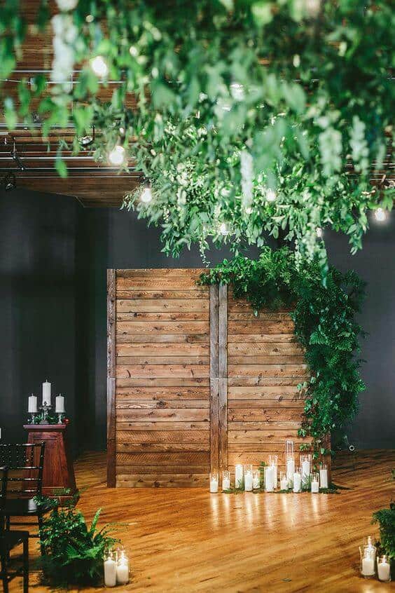 Rustic indoor wedding venue featuring a wooden plank backdrop draped in greenery, glowing candles, and a canopy of hanging foliage.