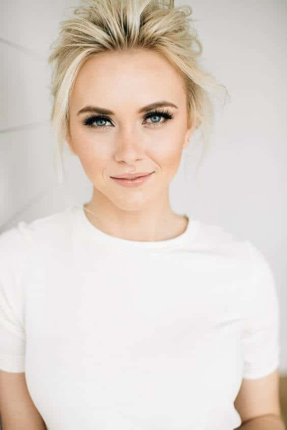 Close-up portrait of a woman with a textured blonde updo and soft glam makeup, wearing a white top against a bright background.