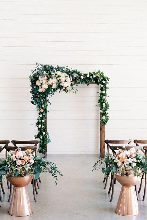 Romantic indoor ceremony featuring a copper-framed wedding arch adorned with lush greenery and pastel florals, set against a bright backdrop.