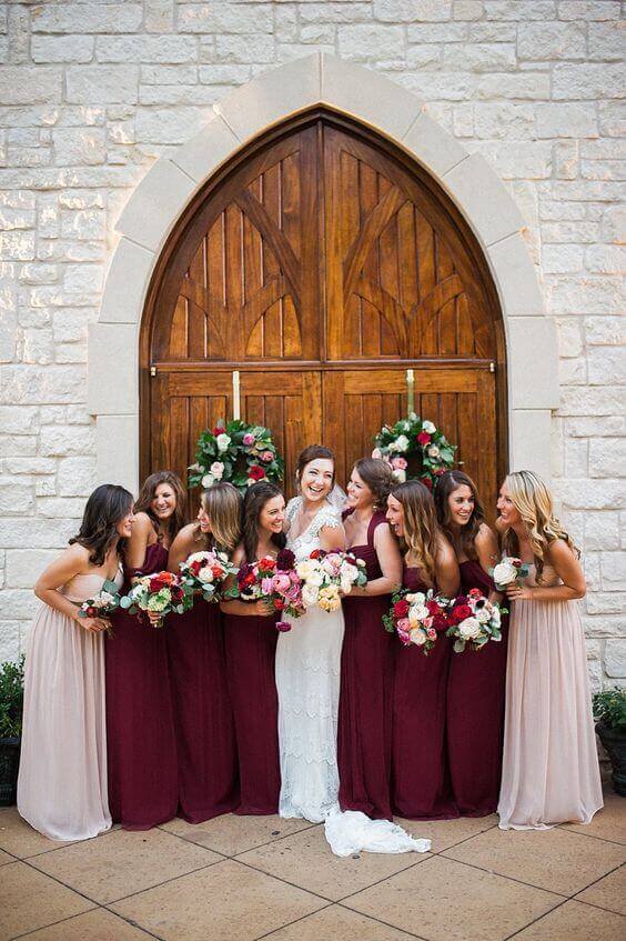 Bridesmaid dress ideas featuring a bride in a lace gown and eight bridesmaids in burgundy and blush at a grand chapel doorway.