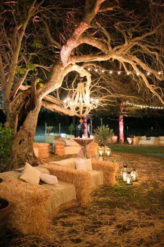 34 budget wedding ideas for a creative affordable wedding 12 Rustic nighttime wedding lounge under a grand tree with bistro lights, hay-bale sofas, and vintage lanterns, creating a cozy ambiance.