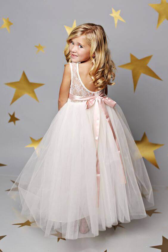 43 cute and perfect flower girl dresses that make every little princess shine 12 A young flower girl in a blush tulle dress with a satin bow gazes over her shoulder, surrounded by metallic gold stars on a dove-gray backdrop.
